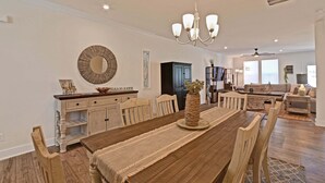 A spacious dining area with a wooden table, chairs, and a chandelier. There is a buffet counter with a mirror and decorative items. The room has a modern ceiling with recessed lighting and a large window providing natural light.