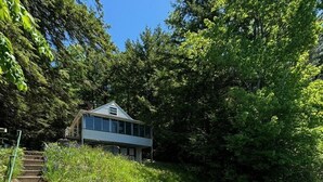 A modern house with a large glass window, surrounded by lush greenery and a stone pathway leading to a body of water.