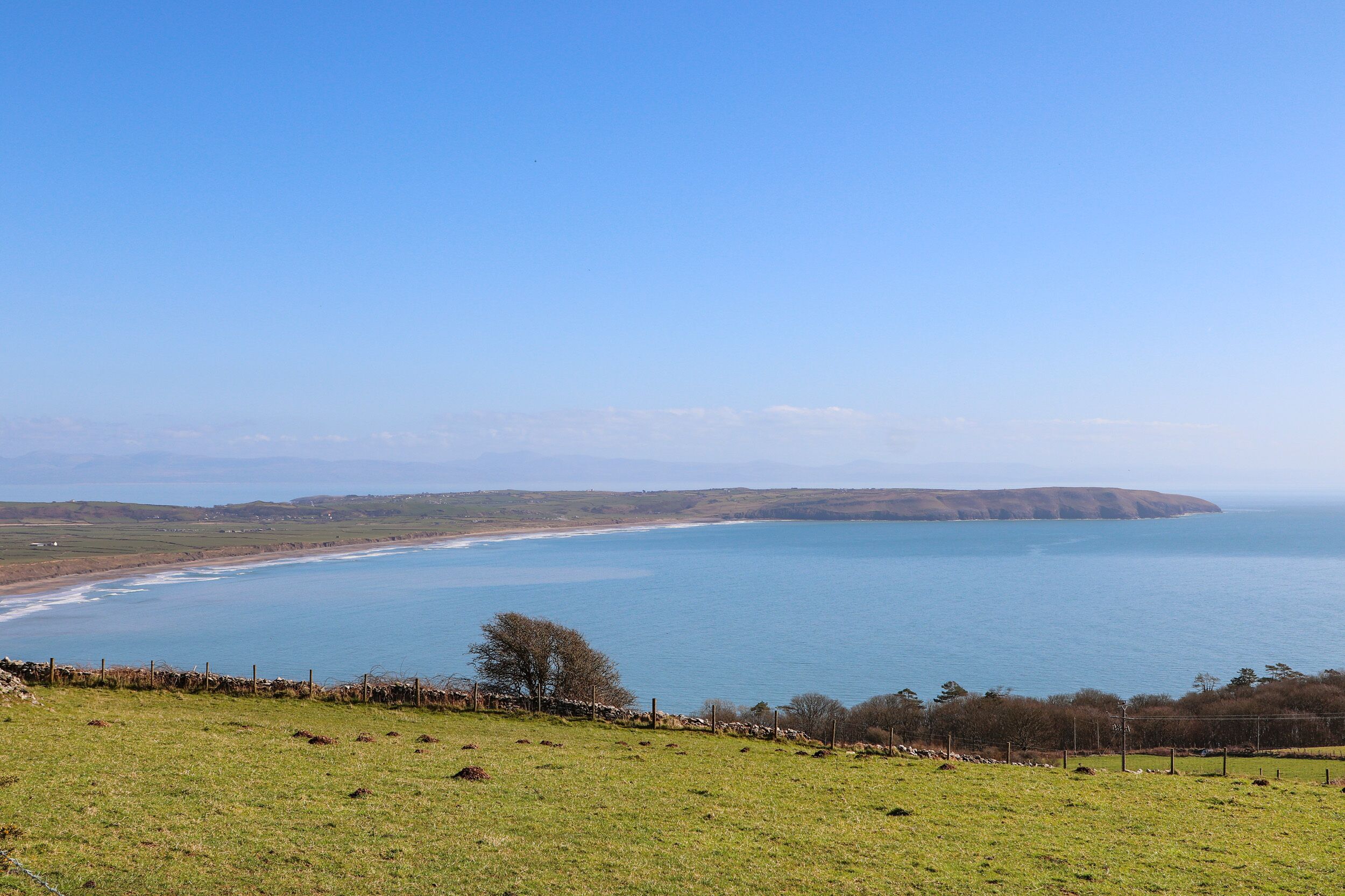 Un paysage côtier avec un champ herbeux, un mur de pierre et une vue sur la mer et les collines lointaines.
