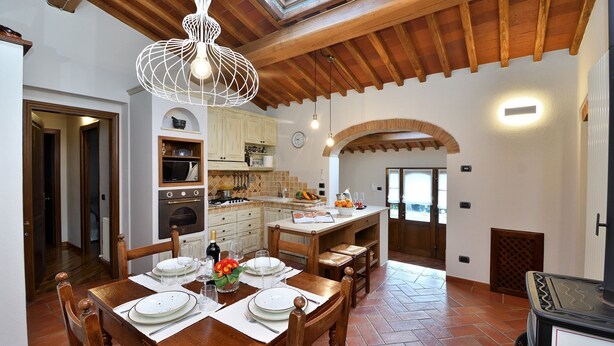 A spacious kitchen with wooden beams, a dining area with a table set for four, and a view of the outdoors through an archway.