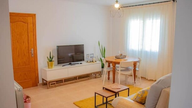 Een moderne woonkamer met een bank, een salontafel, een televisie op een witte kast, een eettafel met stoelen en een raam met gordijnen.