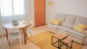 Een woonkamer met een ronde houten tafel, witte stoelen, een bank met decoratieve kussens, en een bijzettafeltje.
