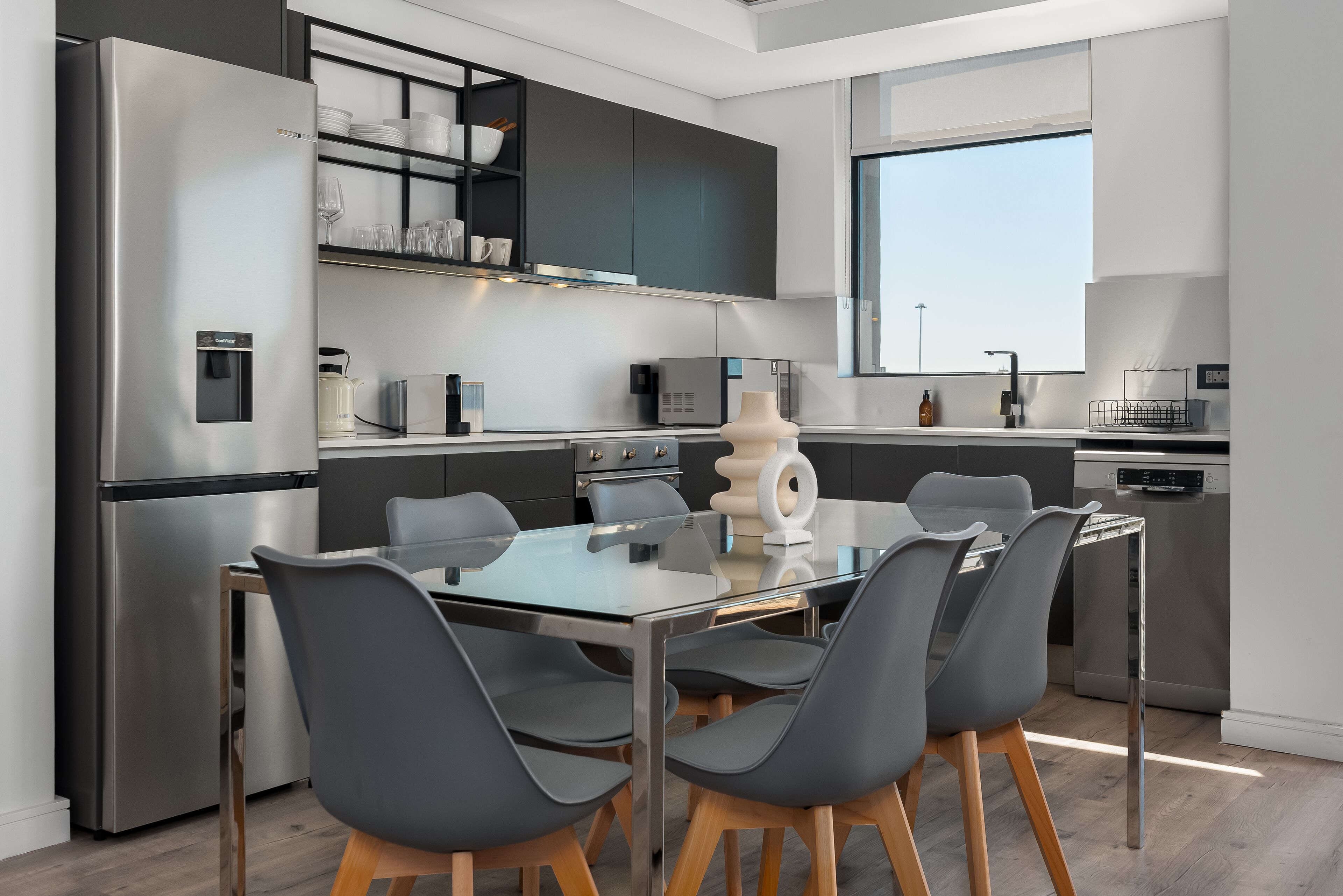 A modern kitchen with a glass dining table, grey chairs, stainless steel appliances, and a black backsplash.