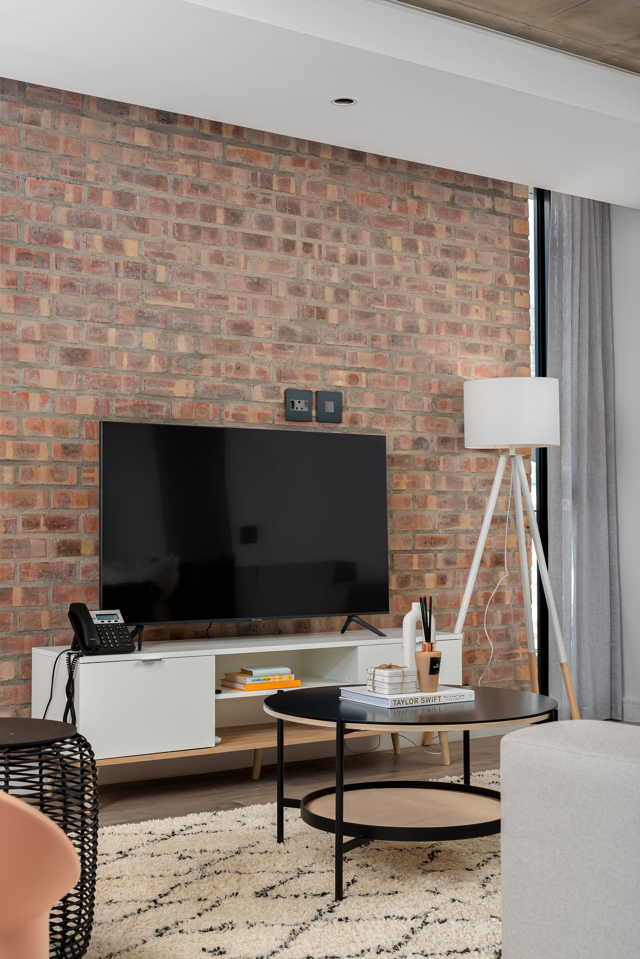 Un salon moderne avec un mur en briques, un téléviseur à écran plat, un meuble de rangement blanc, un lampadaire et une table basse ronde.