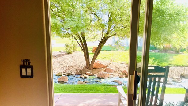 Blick durch eine Glastür auf eine Terrasse mit einem kleinen Bach, einem Baum und einer Holzterrasse mit Stühlen.