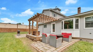 A patio with wicker furniture and a pergola, a house with a chimney, and a wooden fence in the background. - Walk To Wells Harbor! Ping Pong Table & Large Yard (Wells)