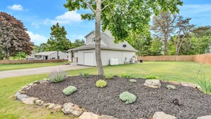 A residential property with a well-maintained lawn, a stone border around the garden, and a house with a gray roof and white siding. - Walk To Wells Harbor! Ping Pong Table & Large Yard (Wells)