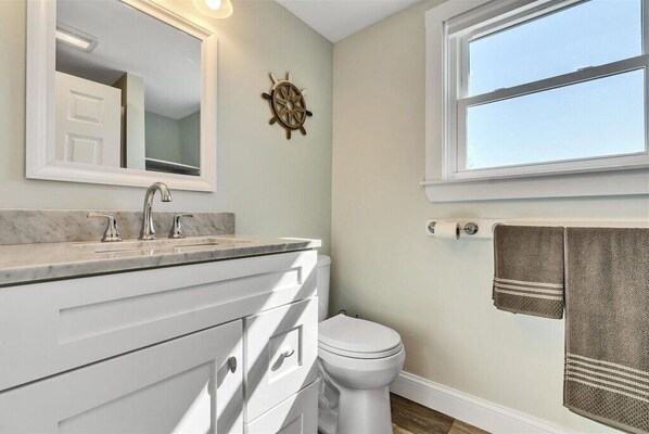 Une salle de bain avec un meuble-lavabo blanc, une décoration en forme de roue de navire, des toilettes et une fenêtre sur laquelle est accrochée une serviette.