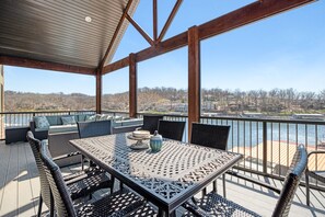 A covered outdoor deck with a table and chairs, overlooking a body of water and a residential area. - Luxury Home on Cove with Boat Slip, Waterslides, and Jump Deck (Linn Creek)