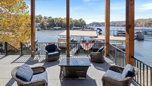 A covered outdoor seating area with wicker furniture and a table, overlooking a lake with boats and docked vessels.