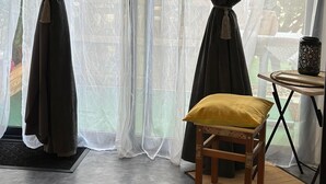 Une pièce avec vue à travers de fines rideaux, un banc en bois avec un coussin jaune et une petite table agrémentée d’un objet décoratif.