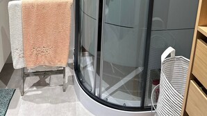 Une salle de bain moderne avec une douche à parois de verre, un porte-serviettes sur lequel est posée une serviette blanche et pêche, et une étagère en bois sur laquelle se trouve un sac à rayures.