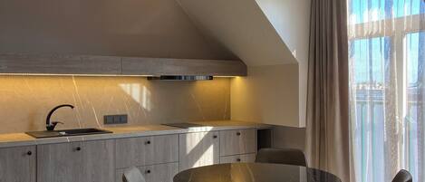 A modern kitchen with a round dining table and two chairs, a built-in oven, and a window with curtains.