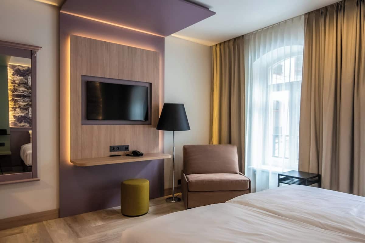 A modern hotel room with a large bed, a flat-screen TV mounted on a wooden panel, a standing lamp, and a chair.