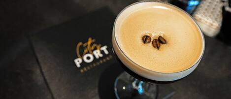 A cocktail with coffee beans on top, served in a stemmed glass, on a bar counter with a 'PORT' sign in the background.