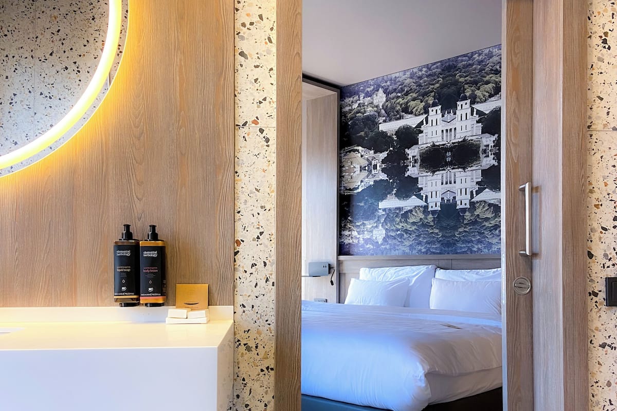 A hotel room with a wooden door, a mirror with a circular light, a bed with white bedding, and a wall-mounted artwork.