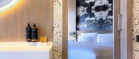 Habitación de hotel con puerta de madera, espejo con luz circular, cama con ropa blanca y una obra de arte colgada en la pared.