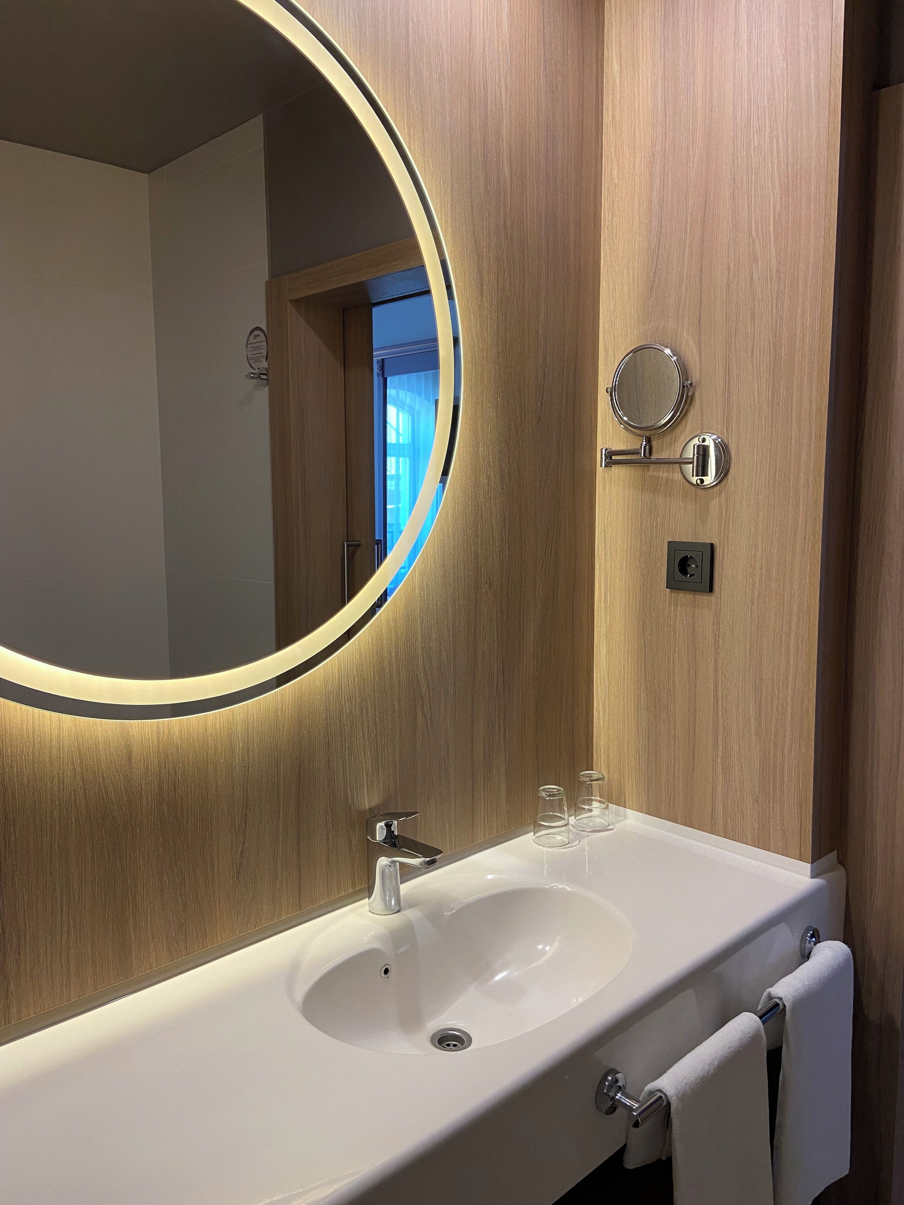 A bathroom with a wooden vanity, a round mirror with a light, a sink, and a towel rack.