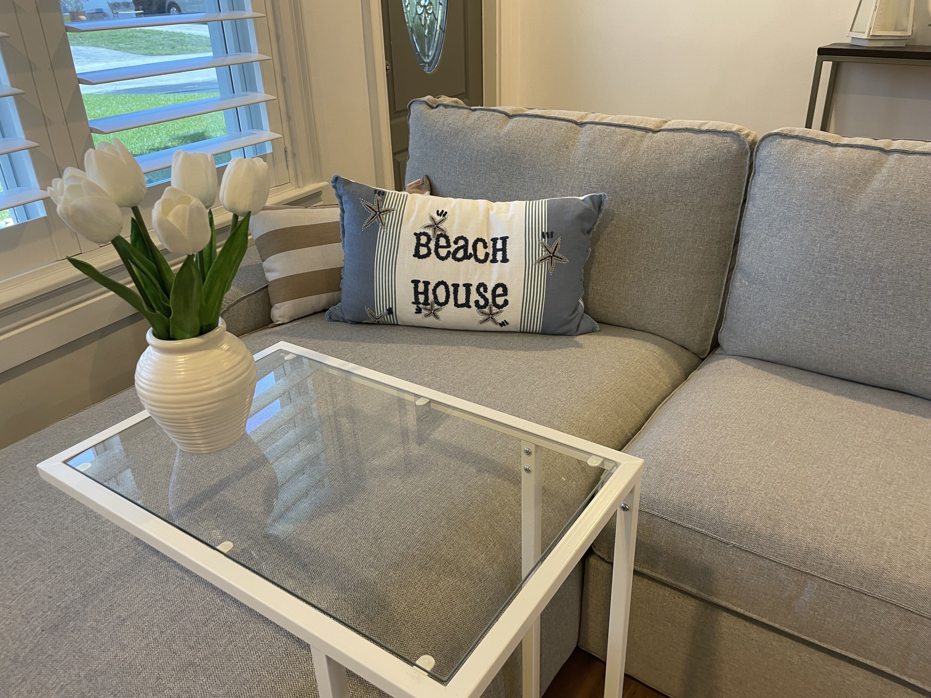 A living room with a glass-top coffee table, a beige sofa, and a decorative pillow with the text "Beach House".