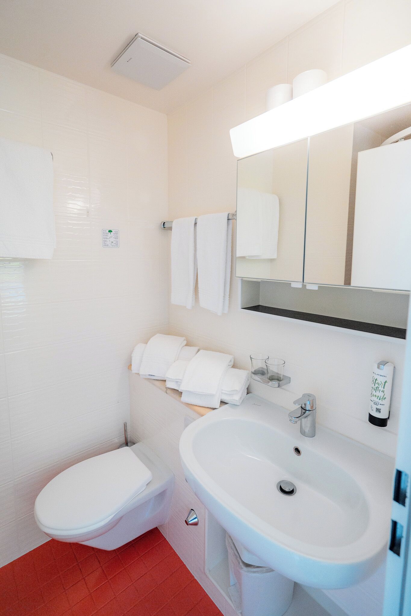 A bathroom with a toilet, sink, and mirror. There are towels and a soap dispenser on the sink counter.