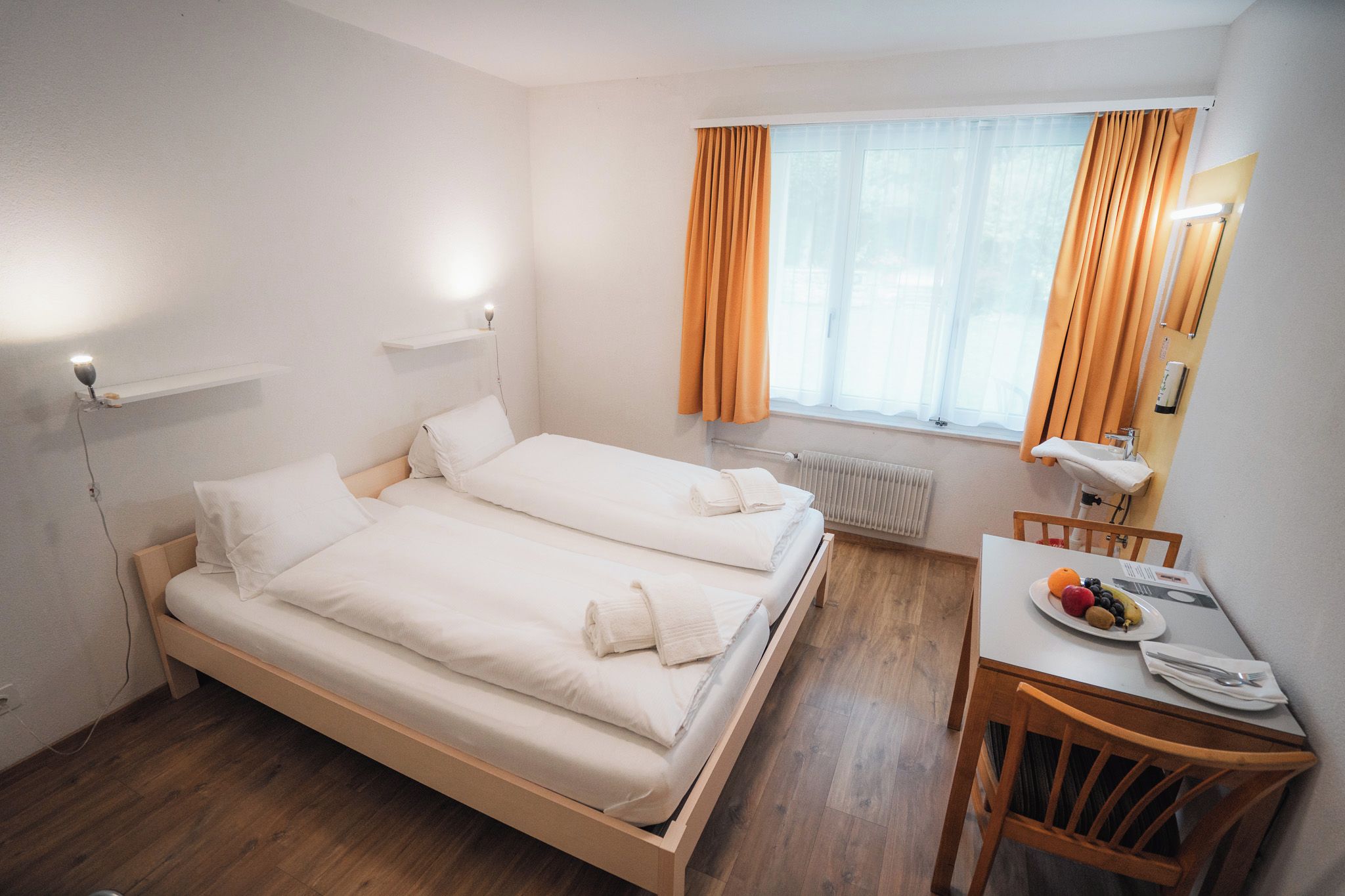 Ein Hotelzimmer mit zwei Betten, einem kleinen Tisch mit einer Fruchtschale, einem Stuhl und einem Fenster mit Vorhängen.