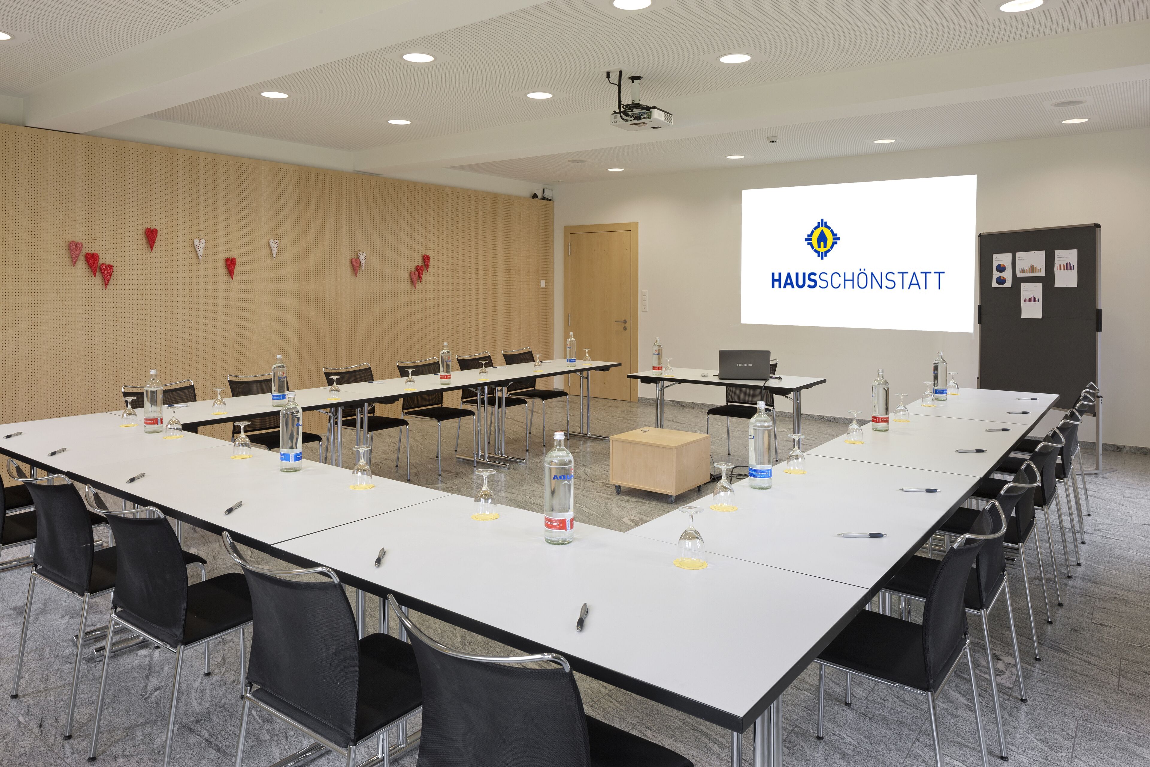 A conference room with a long table, chairs, and a projector screen displaying "HAUSSCHÖNSTATT".
