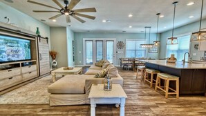 A spacious living room with a large TV, a ceiling fan, and a kitchen area with bar stools.