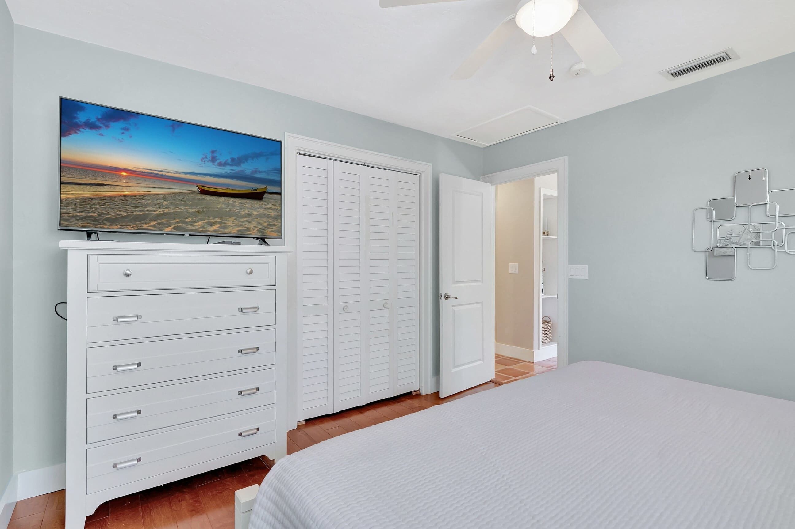 A bedroom with a bed, a dresser with drawers, a closet with doors, a television on the wall, and a ceiling fan.