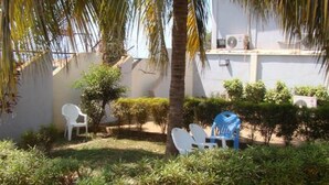Zona d'estar a l'exterior amb cadires de plàstic blanc i un parasol blau, envoltada de vegetació i una palmera.