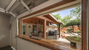 A modern kitchen with a wooden countertop and a sliding glass door leading to a patio area with outdoor seating and a barbecue grill.