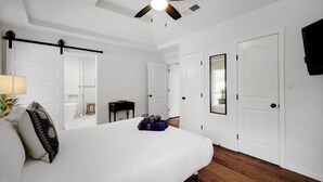 A bedroom with a bed, a ceiling fan, white walls, wooden flooring, and a mounted TV.