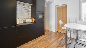 Une cuisine moderne avec des meubles de couleur sombre, un pare-feu en carreaux blancs, un plan de travail en bois, une table à manger blanche avec une chaise, et une porte en bois.