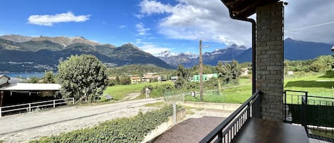 A balcony with a view of a mountainous landscape, a paved area with greenery, and a building with a stone pillar.