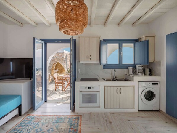 A compact kitchen with white cabinets, a built-in washing machine, and a dining area with a wicker chair.