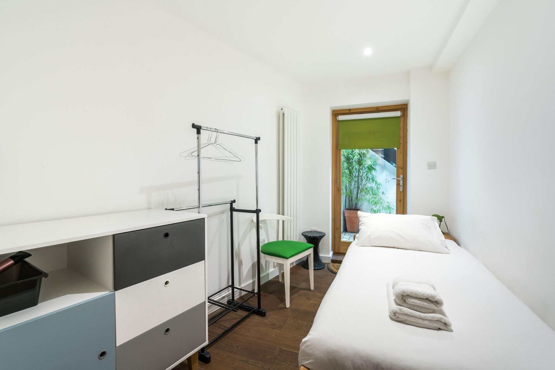 A bedroom with a bed, a desk with a chair, a radiator, and a window with a view of greenery.