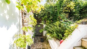 A narrow outdoor space with a brick pathway leading to a small garden area, surrounded by dense foliage and a white wall. - Charming 2 Bedroom Apartment in Kentish Town (London)