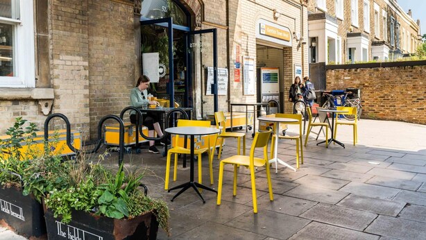 An outdoor seating area with yellow chairs and tables, a brick building with a sign reading "Kentish Town West," and a person sitting at a table. - Charming 2 Bedroom Apartment in Kentish Town (London)