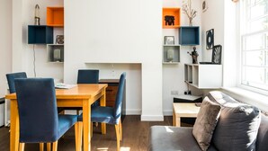 A dining area with a wooden table and blue chairs, a sofa with cushions, and built-in shelving with various decorative items. - Charming 2 Bedroom Apartment in Kentish Town (London)