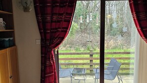 A living room with a black leather sofa, a wooden coffee table, and a sliding glass door leading to an outdoor patio with chairs and a table. - Large 3 bed bungalow set in Woodland grounds near Tamar lakes and local beaches (Kilkhampton)