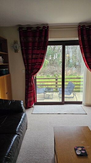 A living room with a black leather sofa, a wooden coffee table, and a sliding glass door leading to an outdoor patio with chairs and a table. - Large 3 bed bungalow set in Woodland grounds near Tamar lakes and local beaches (Kilkhampton)