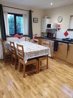 Un comedor con una mesa lista para cuatro personas, sillas de madera, una ventana con cortinas y una cocina con microondas y estufa.