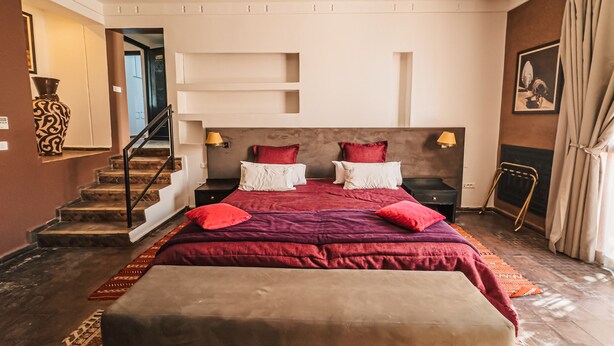 A bedroom with a large bed, two bedside tables, a headboard, a nightstand, a chair, a staircase, and a door leading to another room. - Kasbah Agounsane (Ourika)