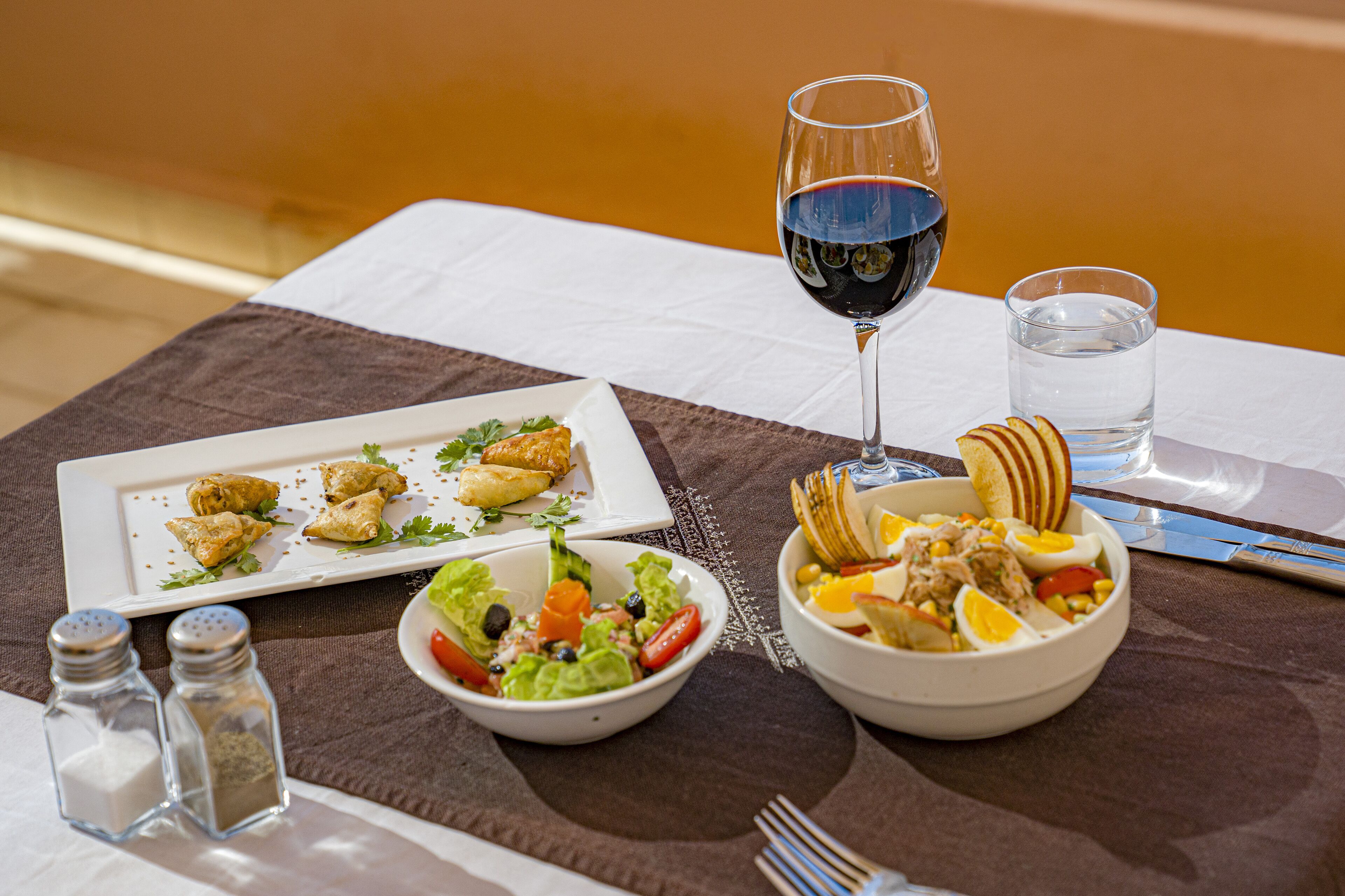 Una mesa servida con un plato de aperitivos, un bol de ensalada, un bol de sopa, una copa de vino tinto y una copa de agua.