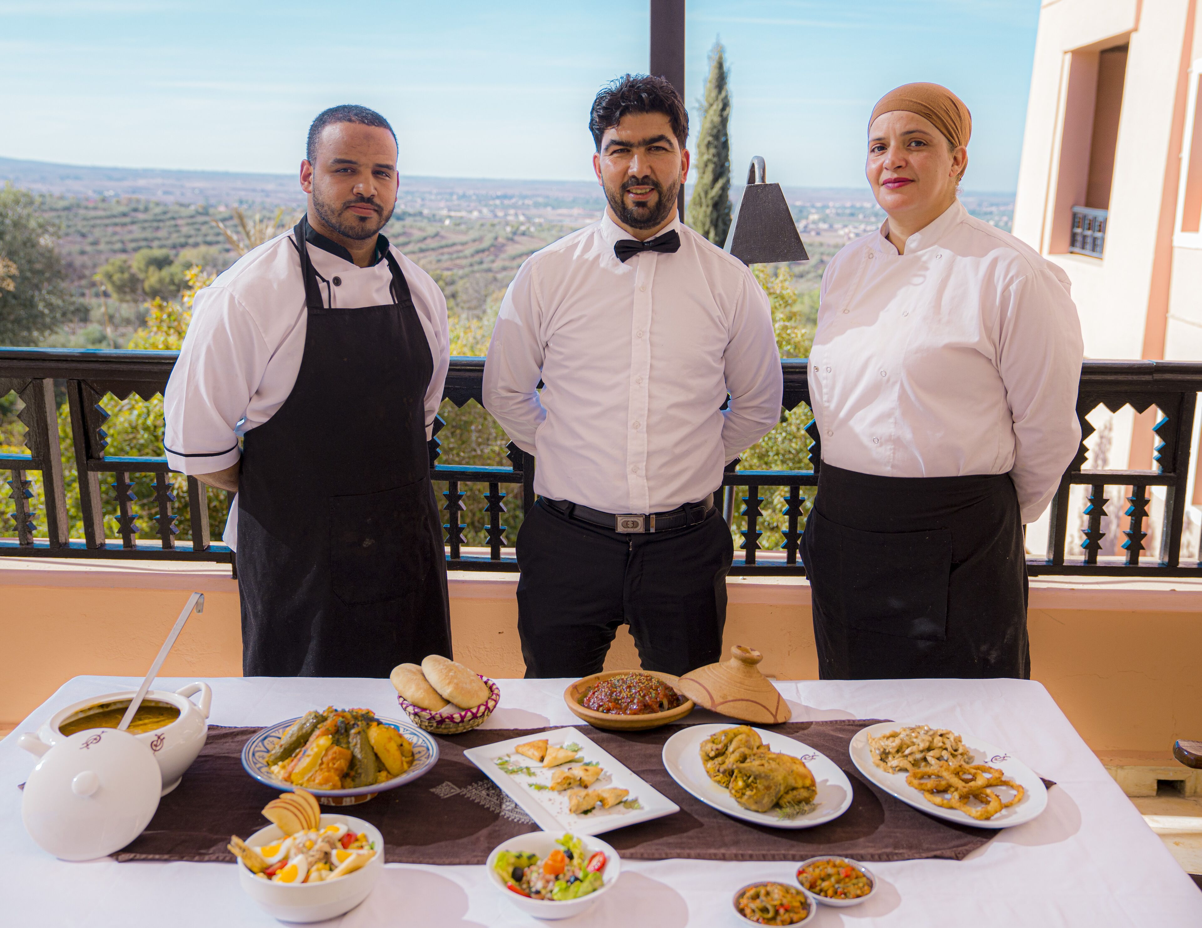 Three waitstaff standing in front of a table with various dishes, a view of the countryside, and a balcony railing.