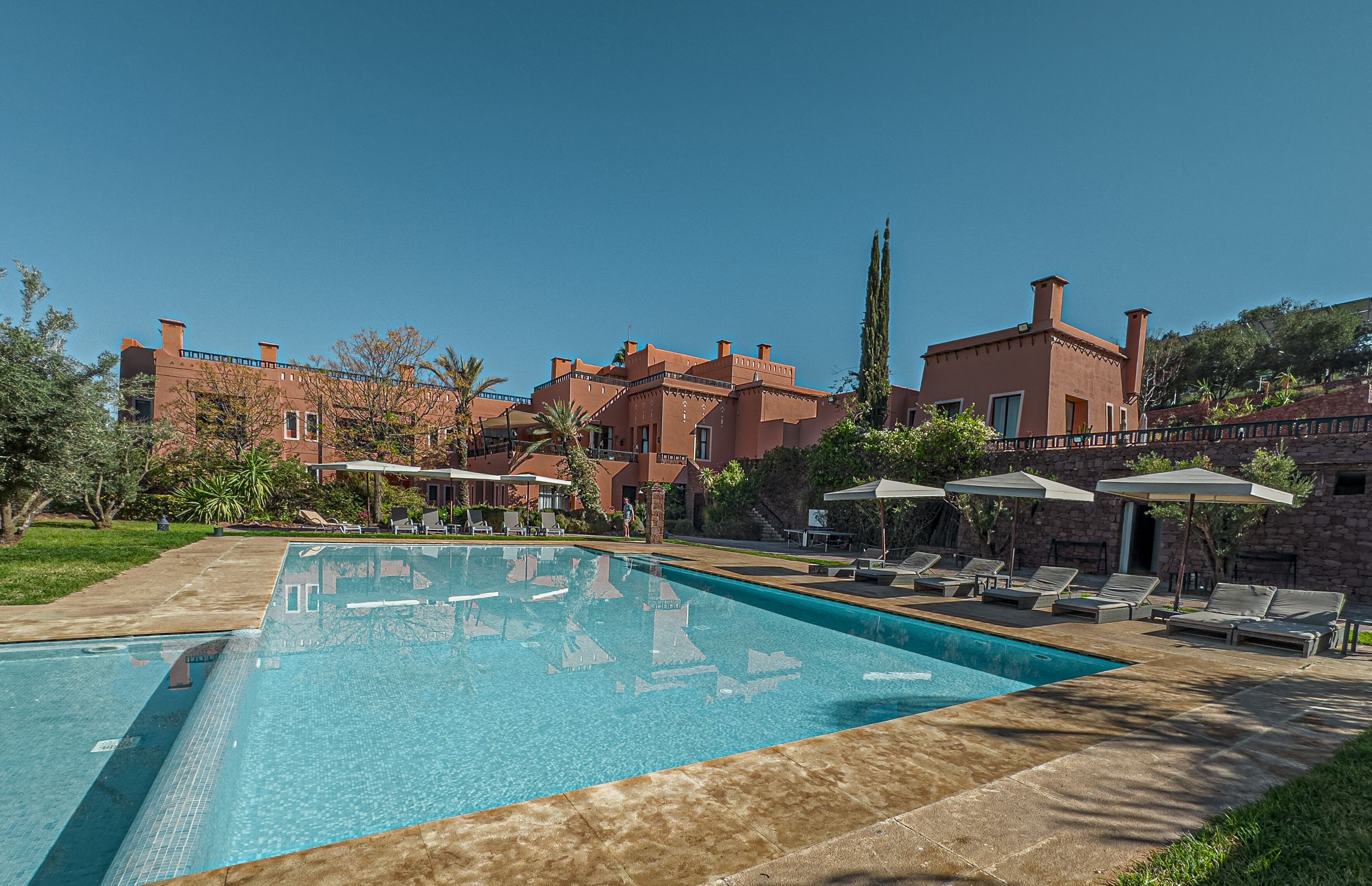 Una piscina con sillas de descanso y sombrillas, rodeada por un edificio de ladrillo con varias chimeneas y un cielo azul despejado.