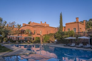 Zona de piscina con sillas de descanso y sombrillas, rodeada por un edificio grande con varios balcones y un árbol alto.
