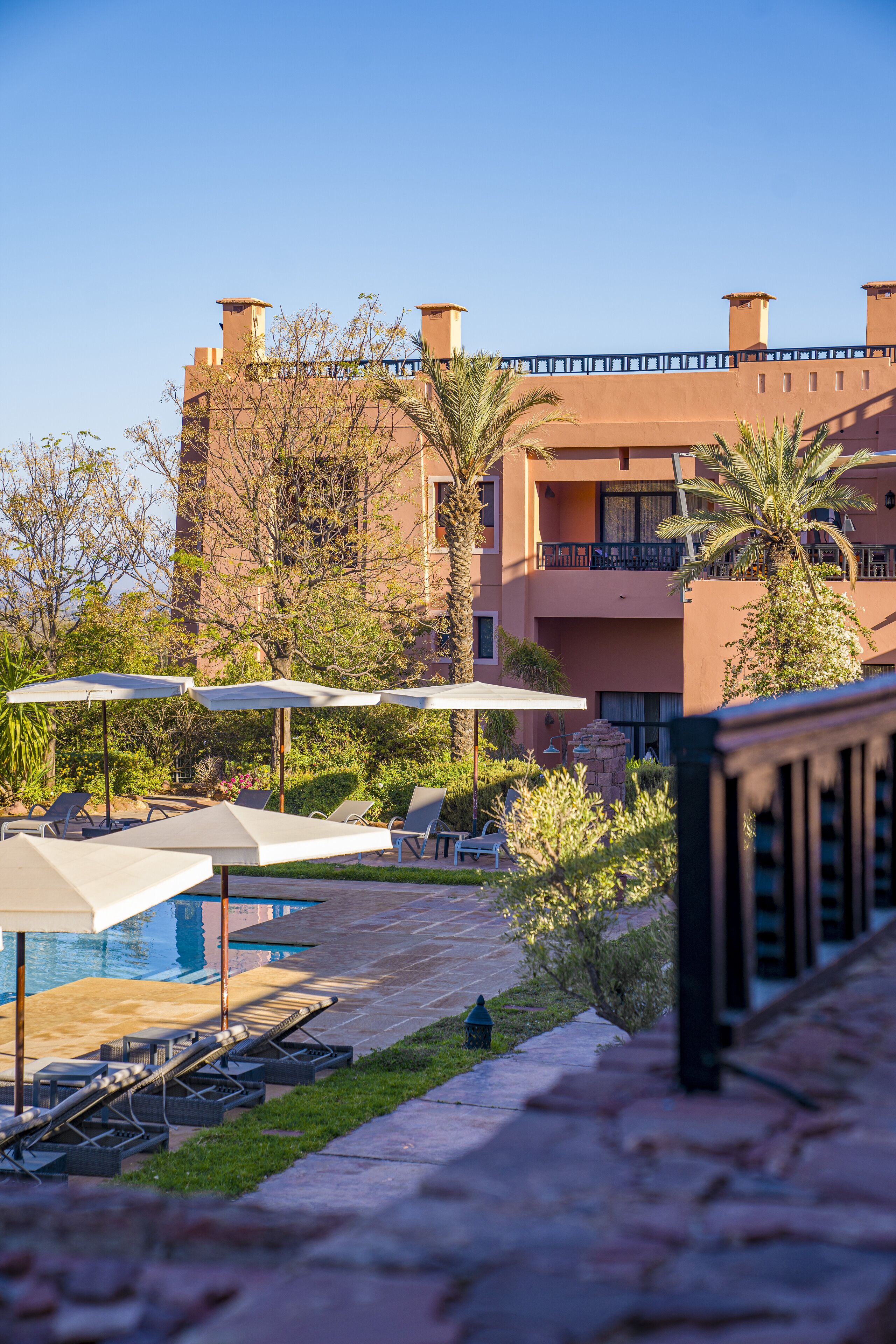 A poolside area with lounge chairs and umbrellas, a clear sky, and a building with balconies and palm trees in the background.