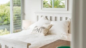 Una cama con ropa de cama blanca, una ventana con persianas y un cuadro colgado sobre la cama.