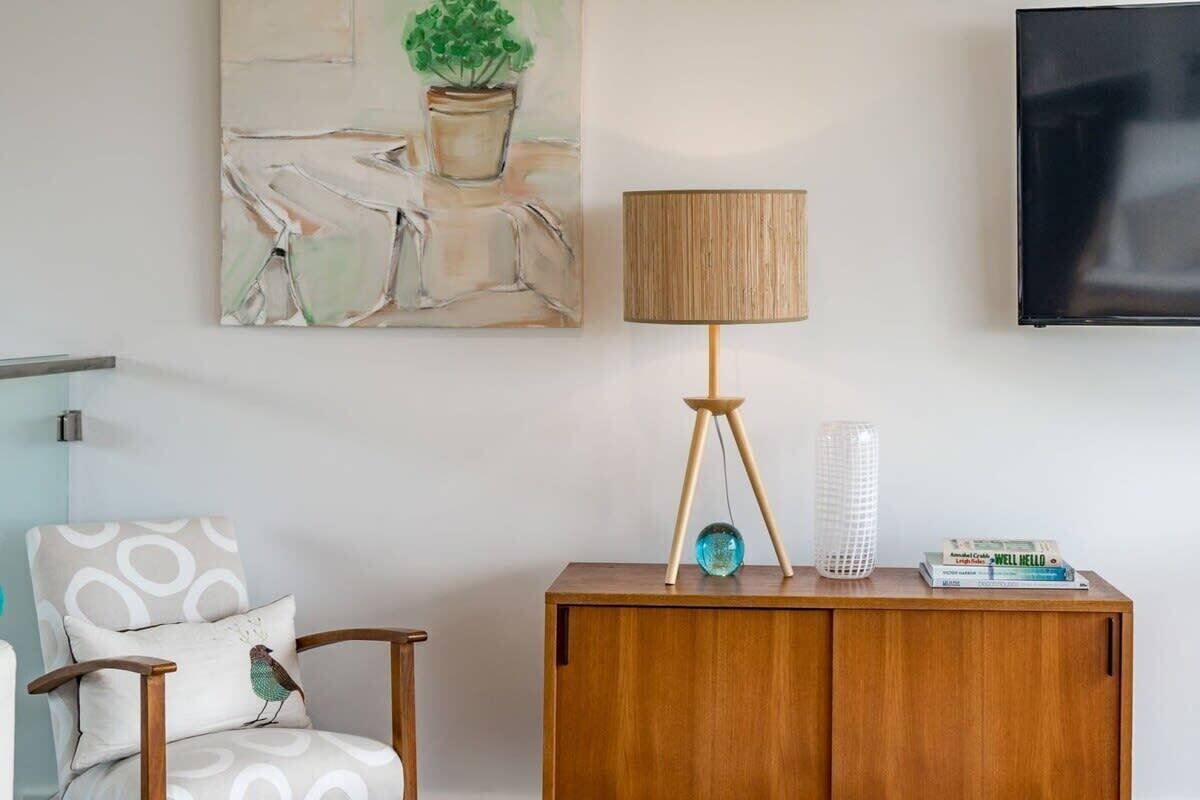 A living room with a wooden cabinet, a lamp, a chair with a cushion, a flat-screen TV, and a painting on the wall.