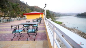 A riverside terrace with outdoor seating, a small building with a red roof, and a view of the river. - Ashoka Resort Pvt. Ltd (Kabilas)